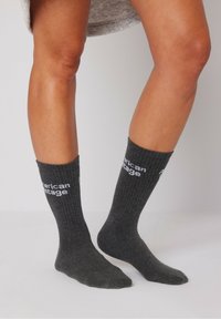 Pilkos medvilnės kulkšnės kojinės su riliniu tekstūra; viršutinėje dalyje yra balti užrašai "American Vintage". Minkštas, kasdieniškas dizainas.