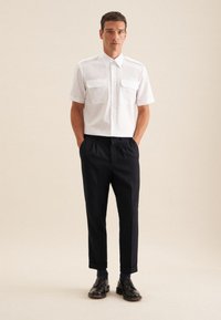 Witte korte mouwen shirt met twee borstzakken, gecombineerd met zwarte high-waisted broeken en zwarte schoenen, tegen een neutrale achtergrond.