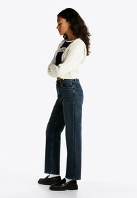 Jeans de tiro alto de color azul oscuro con pierna recta, combinados con un suéter corto de punto trenzado blanco y mocasines negros.