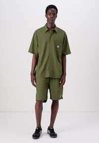 Chemise à manches courtes vert olive et shorts assortis, en tissu texturé, fermeture à boutons, poche avant avec étiquette. Des baskets noires complètent la tenue.