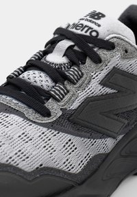 Baskets de sport grises et noires avec des panneaux en mesh et texturés, œillets renforcés et lacets noirs. Présente un logo "N" bien en vue.