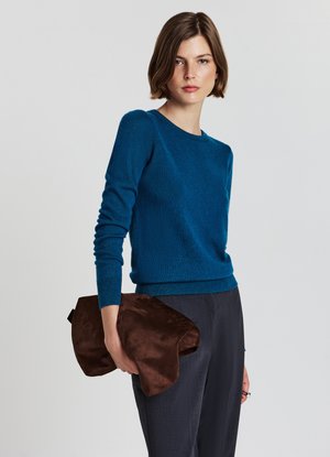 Blue knit sweater with long sleeves and a round neckline. The model holds a brown suede clutch bag. Gray textured trousers complete the outfit.