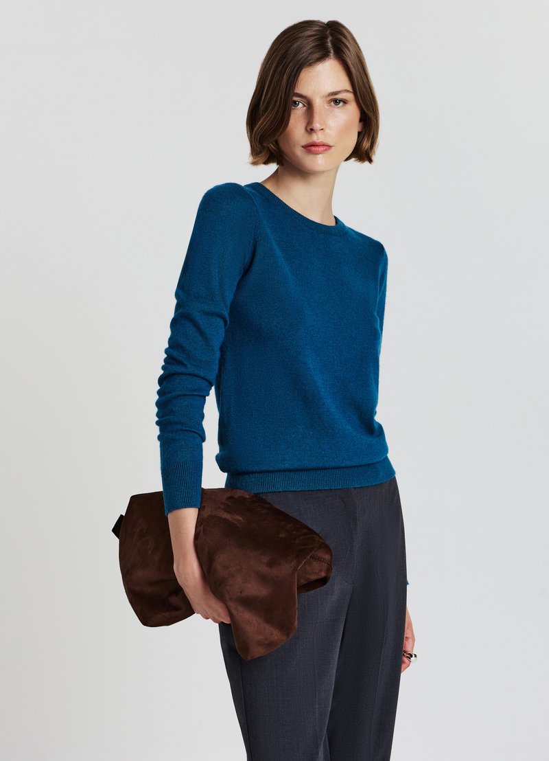 Blauer Strickpullover mit langen Ärmeln und rundem Halsausschnitt. Das Model hält eine braune Wildleder-Clutch. Graue strukturierte Hosen runden das Outfit ab.