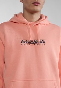 Sudadera color rosa coral fabricada con tejido suave, presenta un bolsillo canguro y un logo impreso en negro que dice "NAPAPIJRI geographic" en el frontal.