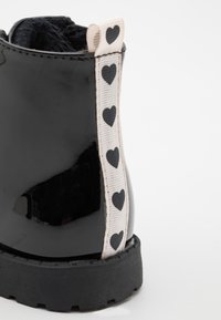 Černá lesklá kožená bota s texturovanou podrážkou, s krémovou stuhou s černými srdíčky po straně.