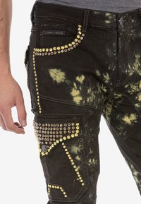 Czarne spodnie w stylu tie-dye z ozdobami ze złotych i brązowych ćwieków na kieszeniach i szwach, noszone przez osobę z widoczną lewą ręką.