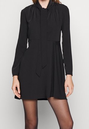 Woman wearing a black long-sleeve mini dress with a tied bow at the neck and black sheer tights, standing against a plain background.