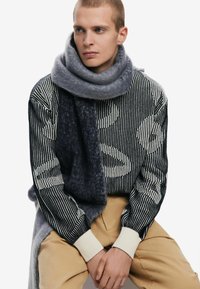 Grå ullsjal som hänger över en randig svart och vit mönstrad tröja med beige byxor. Mjuk textur, förlängd silhuett, kontrasterande färger.