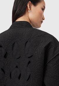 Donna con lunghi capelli scuri indossa una giacca nera trapuntata con design astratti ritagliati sul retro e piccoli orecchini geometrici dorati.