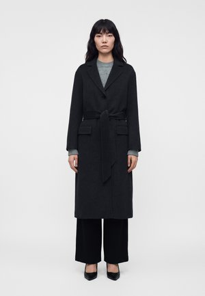 Femme debout portant un long manteau noir ceinturé, un pull gris, un pantalon noir à jambes larges et des chaussures noires à bout pointu sur un fond uni.