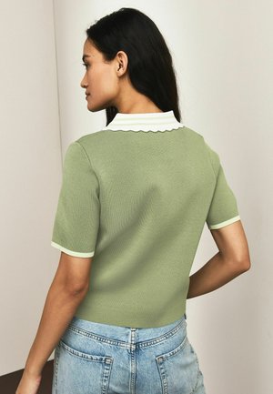 Woman with long dark hair wearing a light green short-sleeve sweater with a white scalloped collar and light blue jeans, facing away.