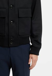 Giacca nera con bottoni, caratterizzata da due tasche sul petto, polsini a coste e un materiale testurizzato simile alla lana. Abbinata a pantaloni blu navy.