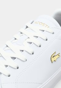 Basket Lacoste blanche avec des lacets blancs, texte Lacoste doré sur la languette et logo crocodile doré sur le côté.