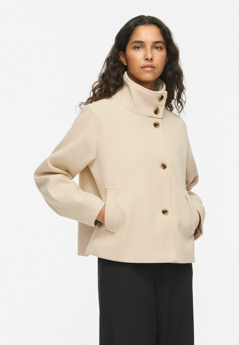 Femme aux longs cheveux bouclés portant une veste courte en laine beige avec un col haut et quatre boutons visibles, mains dans les poches, pantalon noir.