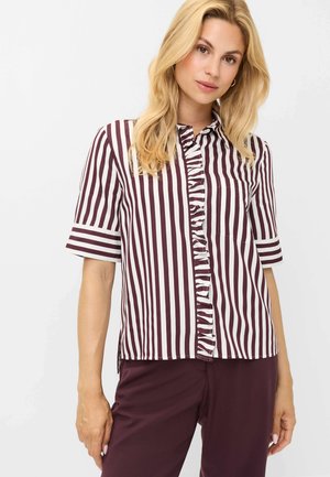Kvinde med blondt hår iført en kortærmet hvid og mørkerød vertikalt striped bluse med flæser foran og mørkerøde bukser.