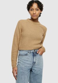 Cropped, textured sweater in tan with a round neckline. Paired with high-waisted faded blue jeans. Model standing against a plain background.