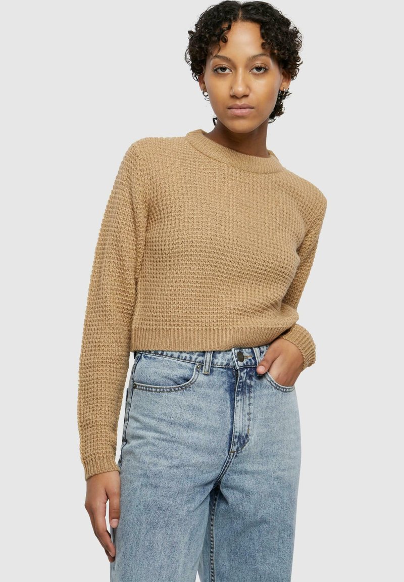 Cropped, textured sweater in tan with a round neckline. Paired with high-waisted faded blue jeans. Model standing against a plain background.