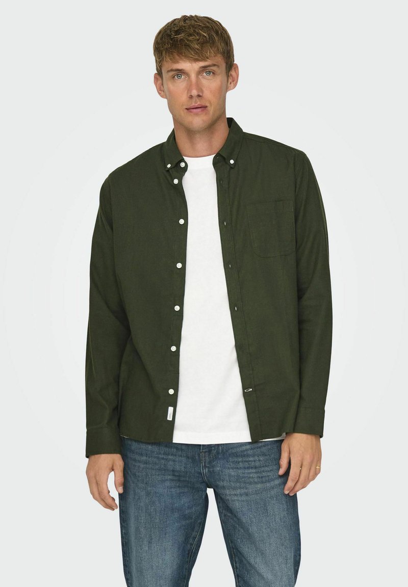 Camisa abotonada verde oscura con mangas largas, un bolsillo en el pecho y botones blancos, utilizada sobre una camiseta blanca y combinada con jeans azules.