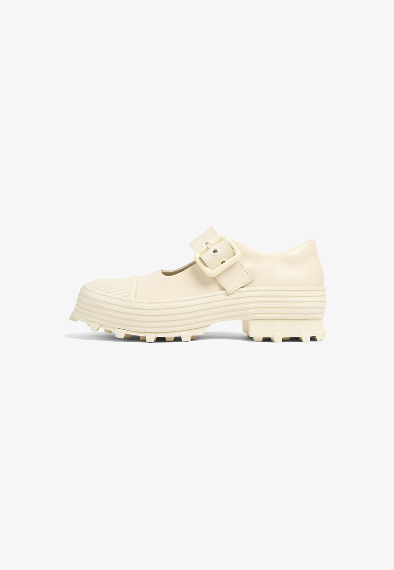 Cream-colored shoe with a rubber outsole, featuring a strap and buckle detail, ridged toe design, and elevated heel with textured grooves.