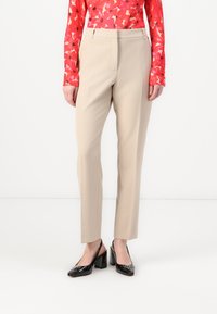 Pantalons beiges ajustés à coupe droite, avec une braguette zippée et des poches latérales, associés à des escarpins en cuir laqué noir.