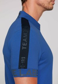 Camiseta polo azul hecha de algodón. Presenta rayas de acento negras en los hombros con el texto "EQUIPO" y el detalle "LA ETIQUETA" en la manga.