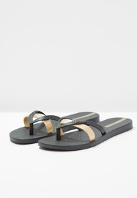 Tongs noires avec une surface texturée, présentant un accent métallique doré sur la lanière et un entre-doigts central. Design léger.