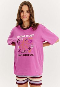 Růžové oversize tričko s vícerozměrným grafickým textem a ilustracemi, spárované s pruhovanými háčkovanými šortkami v různých barvách.