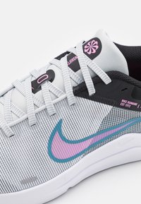 Grå textilsneaker med en ovandel i nät, med en turkos och rosa Nike-swoosh, svarta detaljer och en platt vit sula. Skosnören är ljusgrå.
