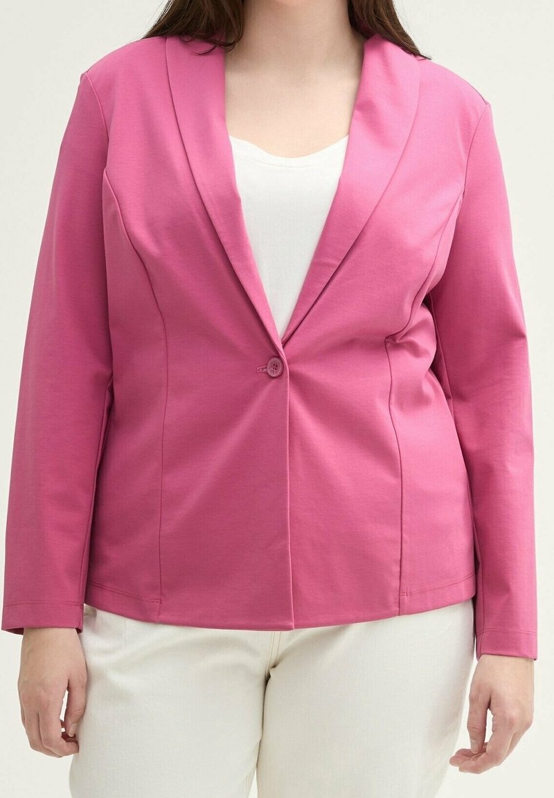 Femme portant un blazer rose vif à un bouton sur un haut blanc et un pantalon beige clair, debout avec les bras détendus le long du corps.