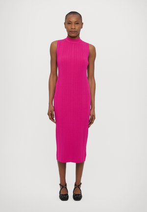 Fitted, ribbed, sleeveless dress in bright pink, with a high neck and knee-length hem. Accents include black, polka-dot shoes.