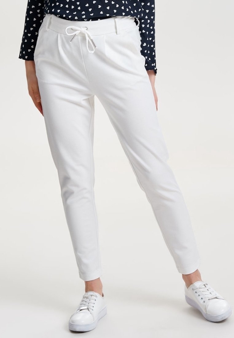 Pantalon slim blanc avec taille à cordon, porté avec des baskets blanches et un top noir avec motifs de cœurs blancs.