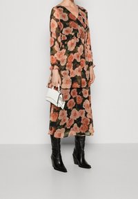 Maxikleid aus schwarzem Chiffon mit pinkem Rosenmuster, mit V-Ausschnitt, elastischem Bund und langen Ärmeln. Kombiniert mit schwarzen kniehohen Stiefeln und einer weißen Handtasche.