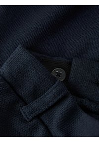 Tejido de punto en color azul marino con superficie texturizada, que presenta un botón negro y una correa en bucle. Acercamiento que resalta los detalles del tejido y la costura.