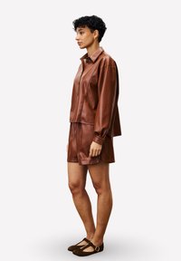 Femme aux cheveux courts et bouclés, portant une veste en cuir marron à boutons et un short assorti, se tenant de profil avec des ballerines marron.