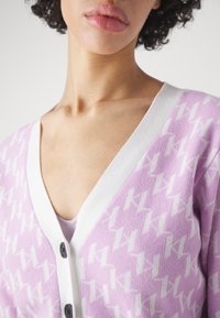 Světlofialový kardigan s bílým geometrickým vzorem, výstřihem do V a bílým lemováním. Má tmavé knoflíky po celé přední straně. Jemná textura.