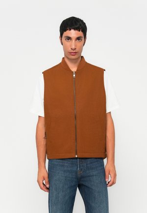 Man wearing brown zip-up sleeveless vest over white t-shirt and blue jeans, standing against plain light background.