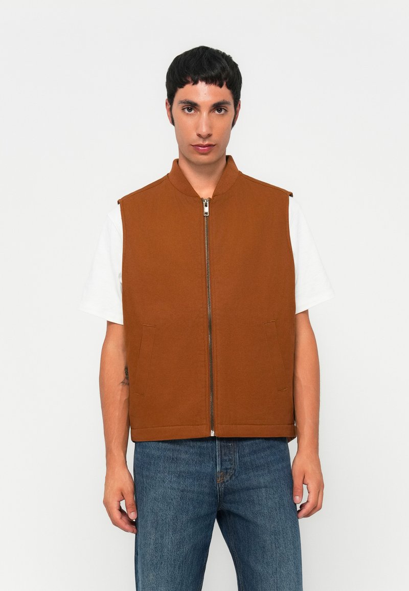 Homme portant un gilet sans manches zippé marron sur un t-shirt blanc et un jean bleu, se tenant contre un fond clair.