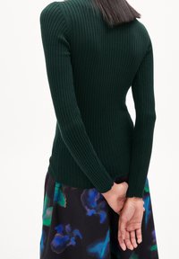 Top a maniche lunghe a coste verde scuro con silhouette aderente, scollo rotondo e tessuto testurizzato; abbinato a una gonna a fantasia floreale colorata.
