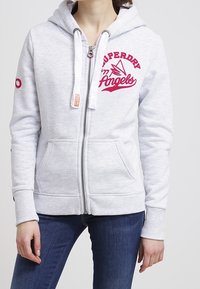 Sudadera con cremallera de color gris claro, con bolsillos frontales, que presenta el logo rojo de Superdry Angels en el pecho y detalles en rosa en las mangas. Textura de tela suave.