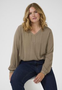 V-veida kakla, garām piedurknēm blouse bālā taupe krāsā, izgatavota no viegla teksturēta auduma, kombinēta ar tumši ziliem džinsiem.