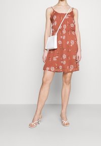 Vestito arancione ruggine con spalline a spaghetti, stampa floreale, vita cintrata e lunghezza sopra il ginocchio. Abbinato a una borsa a tracolla bianca e tacchi con cinturini.