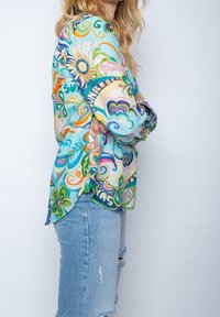 Kvinde med langt blont hår, iført en farverig blomstret skjorte og lyseblå slidte jeans, stående med armene kors. 