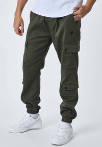 Persona che indossa pantaloni cargo verde oliva con polsini elasticizzati e sneaker bianche, posando con una mano in tasca e l'altra rilassata.