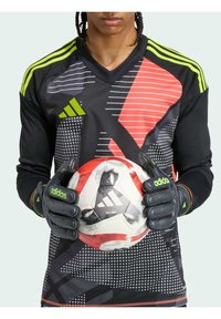 Adidas doelman shirt met zwarte, rode en grijze geometrische patronen, voorzien van neon groene strepen en handschoenen, terwijl hij een voetbal vasthoudt.
