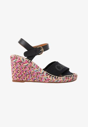 Sandale compensée noire à bride cheville, talon tissé multicolore, bride en tissu au niveau des orteils avec nom embossé et fermeture à boucle dorée.