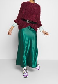 Bordový žebrovaný svetr, zelená saténová maxi sukně, černý pásek a vícebarevné tenisky s detaily, které zahrnují tkaničky a gumové podrážky.