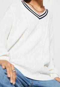 Vit stickad tröja med diamantmönster, v-ringad och med marinblå randig detalj vid kragen. Ärmarna är ribbade. Kombinerad med blå denim.