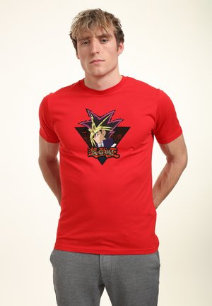 Junger Mann mit blondem Haar, der ein rotes T-Shirt mit einer Figur aus Yu-Gi-Oh vor einem schwarzen dreieckigen Hintergrund und graue Hose trägt.