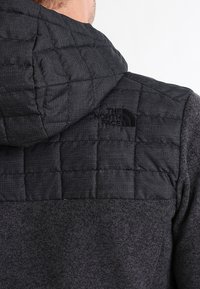 Mies, jolla on päällään tummanharmaa tilkkuhuppari, jossa on The North Face -logo selässä.