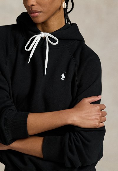 Vrouw die een zwarte hoodie draagt met witte trekkoorden en een klein wit geborduurd logo, met gekruiste armen, en een oorbel in de vorm van een schelp.